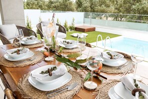 A dining area with a pool view, set tables with white plates, glasses, and wooden utensils, and a wicker chair.