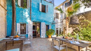 An outdoor dining area with tables set for two, surrounded by a blue building with shutters, potted plants, and a hanging basket. - Casa Metge Vidal by Balearic Villas (Pollensa)