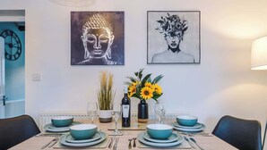 Mesa de comedor decorada con platos, fuentes y cubiertos en color turquesa, flanqueada por sillas negras. Sobre la mesa, hay dos obras de arte enmarcadas, una que representa a Buda y otra a una mujer con flores. En la mesa también hay un jarrón con girasoles y una botella de vino.