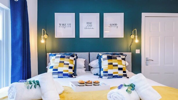 Una cama bien hecha con almohadones a cuadros, una bandeja con muffins y tres carteles motivacionales en la pared.