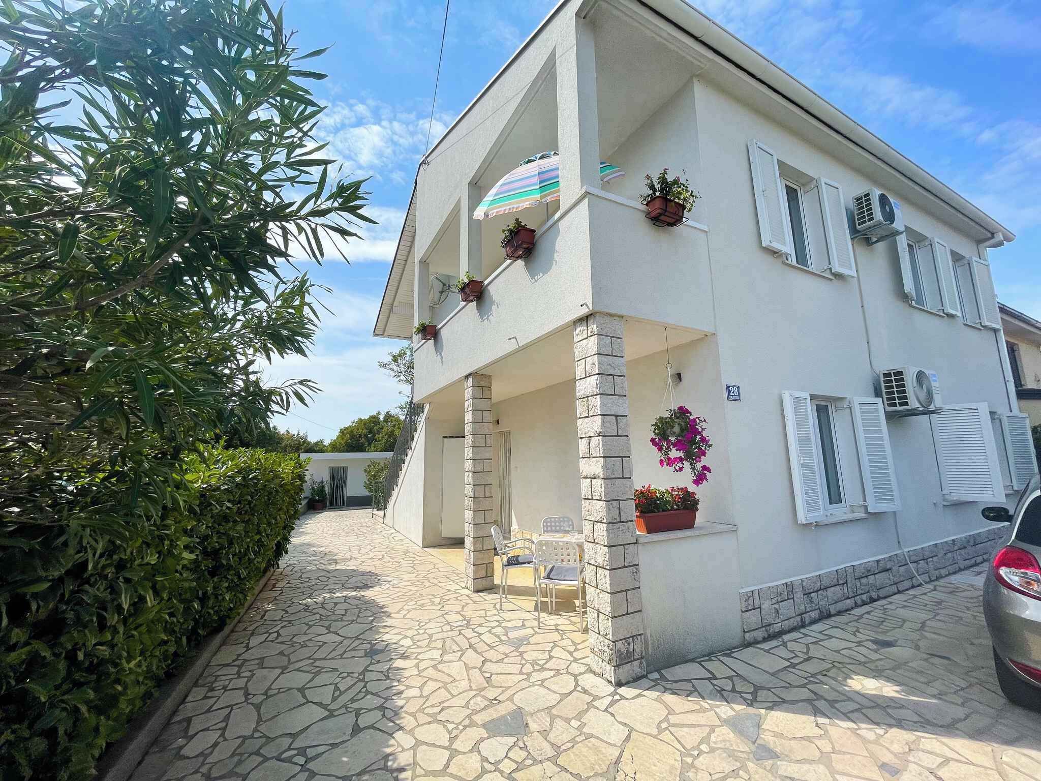 A two-story building with a stone-paved entrance, potted plants, and a striped umbrella on the balcony.