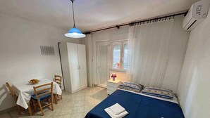 Een kleine, nette slaapkamer met een eenpersoonsbed, een nachtkastje, een stoel, een tafel met een fruitschaal en een raam met gordijnen.