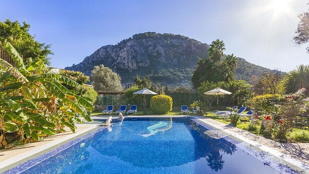 A clear blue swimming pool with lounge chairs and umbrellas, surrounded by greenery and a mountainous backdrop.