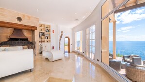Un salón amplio con chimenea, un sofá blanco y vistas al mar a través de grandes ventanales.