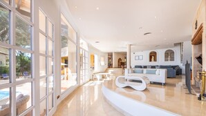 Amplia sala de estar con un sofá blanco moderno, una mesa de centro blanca curva y un gran ventanal con vista a una zona de piscina.