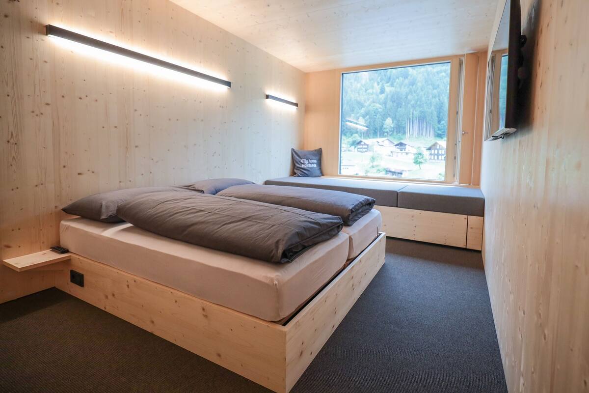 Une chambre dans une cabane en bois, avec un lit, un coin fenêtre aménagé en siège et un téléviseur fixé au mur.