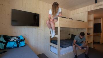 A bunk bed room with a television mounted on the wall, a suitcase on the floor, and a person sitting on the lower bunk.