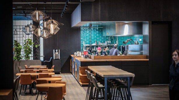 Een modern restaurant met houten tafels en stoelen, een zichtbare keuken door een glazen wand, en hangende pendantlampen.