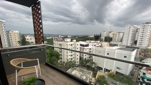 A balcony with wicker chairs and a small table, overlooking a cityscape with various buildings and a cloudy sky.