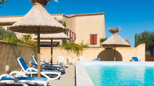Un espace aménagé au bord de la piscine, avec des chaises longues et des parasols en chaume, un ciel d’un bleu éclatant et un bâtiment doté d’un toit en tuiles.