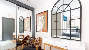 A modern room with a large arched mirror, a wooden chair with a patterned cushion, a white desk, and framed artwork on the wall.