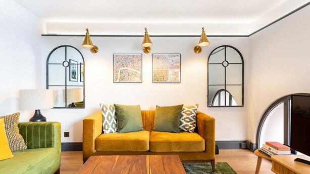 A living room with a green sofa, a mustard-colored sofa, and a wooden coffee table. There are two wall-mounted lamps, a TV, and framed artwork on the wall.