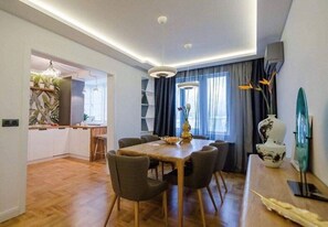 A modern dining area with a wooden table and chairs, a chandelier, and a view of the kitchen and living space. - Clock Apartments (Sveti Vlas)