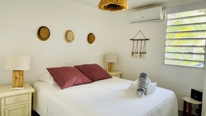Une chambre à coucher comprenant un lit, deux tables de chevet, un luminaire suspendu, un climatiseur fixé au mur et une fenêtre avec des stores.