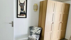 Une pièce avec une porte blanche, un tableau encadré représentant un chien, une armoire en bois, une chaise à motifs tropicaux et une petite lampe fixée au mur.