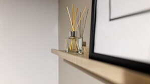 Eine transparente Glasflasche mit Schilfrohrendstäben auf einem Holzregal vor einer schlichten Wand.