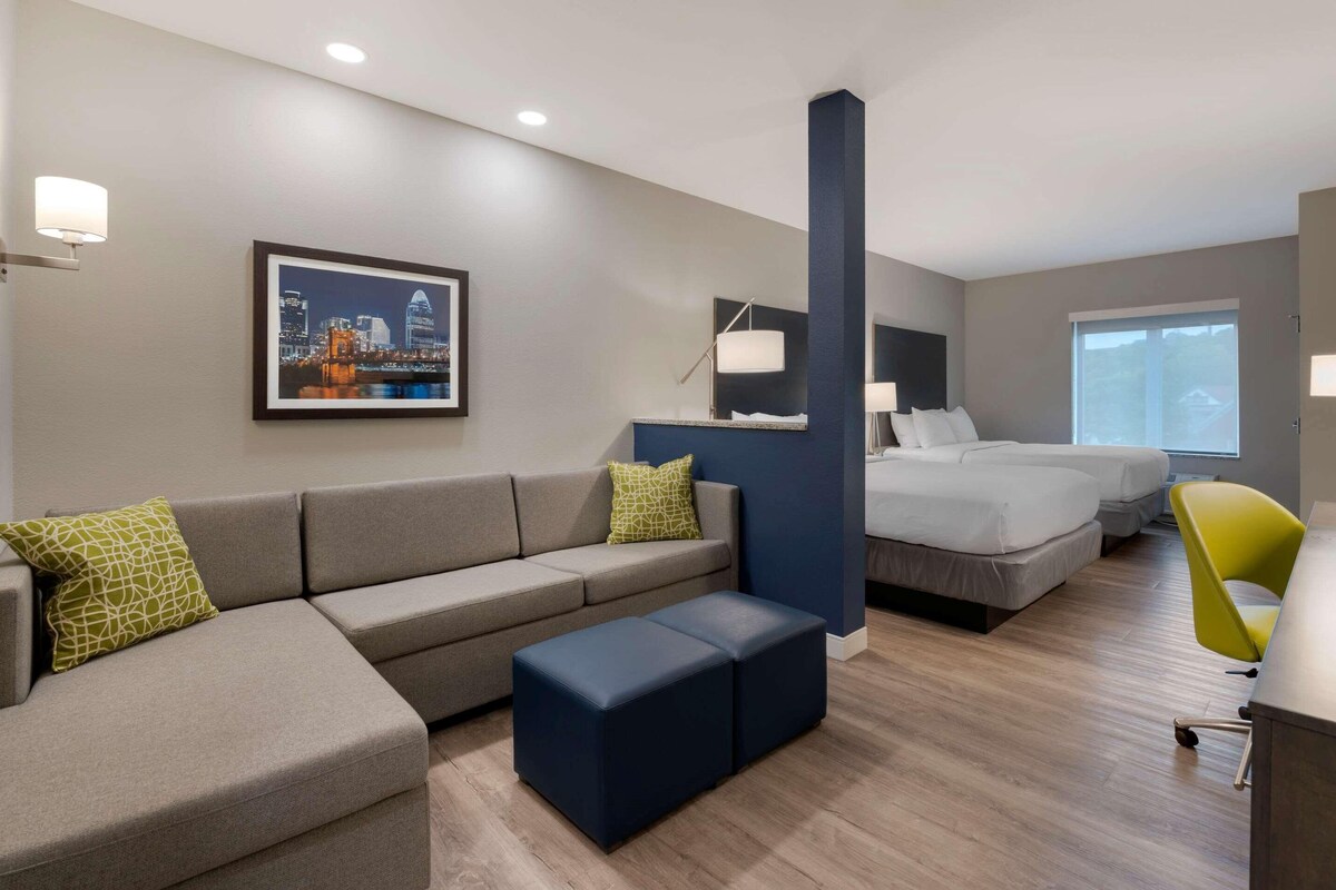 A modern hotel room with a grey sofa, a bed, a desk, and a cityscape painting on the wall.