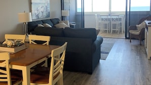A living room with a dark blue sofa, a wooden dining table with chairs, a ceiling fan, and a large window offering a view of the ocean. - Direct Beachfront with pool (Ocean City)