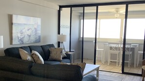 A modern living room with a dark grey sofa, a white coffee table, and a painting of boats on the wall. - Direct Beachfront with pool (Ocean City)