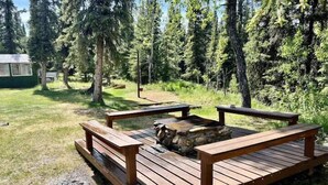 Eine Holzterrasse mit Bänken und einer Feuerstelle aus Stein, umgeben von Bäumen.