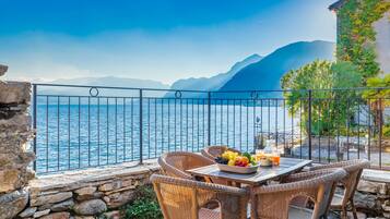 Un balcone con mobili in vimini, un tavolo con frutta e bevande, e vista sul mare e sulle montagne.
