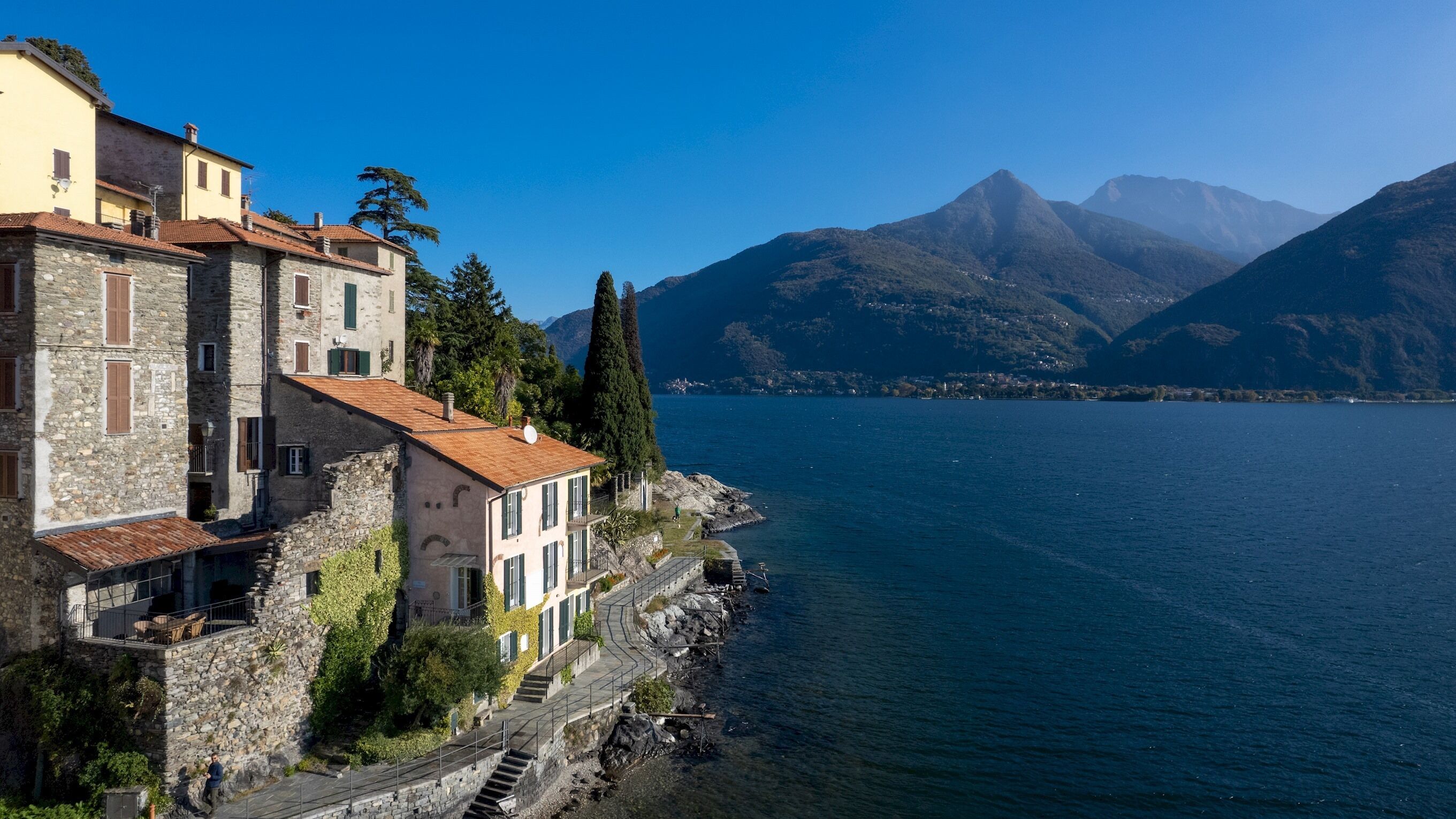 Un edificio costiero con muri in pietra e un tetto in tegole rosse, vicino a un lago calmo con montagne sullo sfondo.