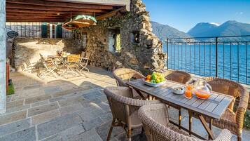 Un patio in pietra con mobili in vimini, un tavolo di legno con frutta e bevande, e vista sul mare e sulle montagne.
