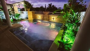 A heated outdoor swimming pool with a hot tub, surrounded by plants and a stone wall with lighting. - Beach Rental - Pet Friendly (chabihau)