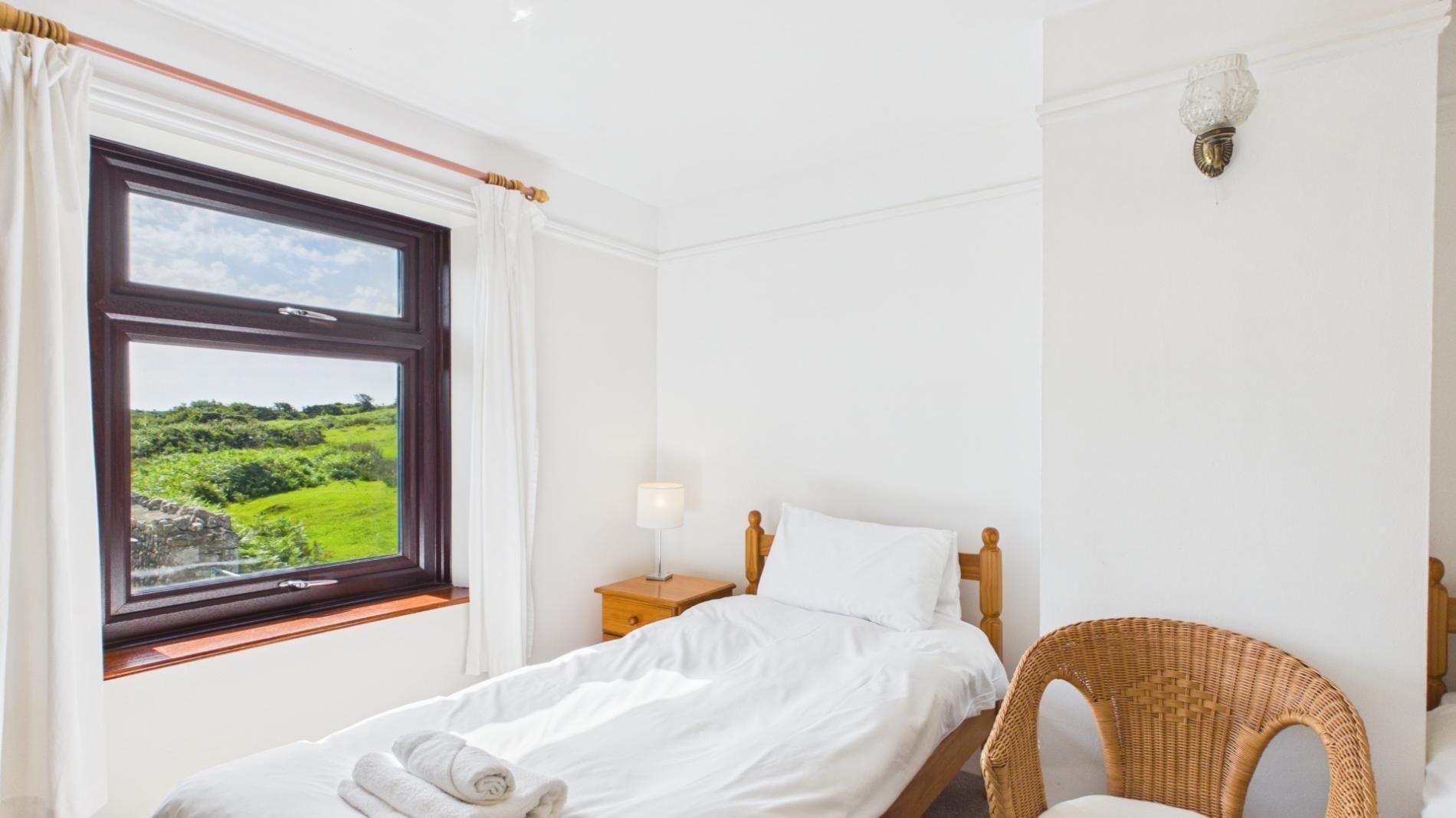 Une chambre à coucher comprenant un lit, une table de chevet, une lampe, un fauteuil en osier et une fenêtre donnant sur un paysage verdoyant.