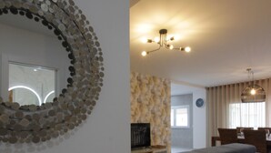 A modern living room with a large round mirror, a wooden side table, a grey sofa, and a dining area with a chandelier.