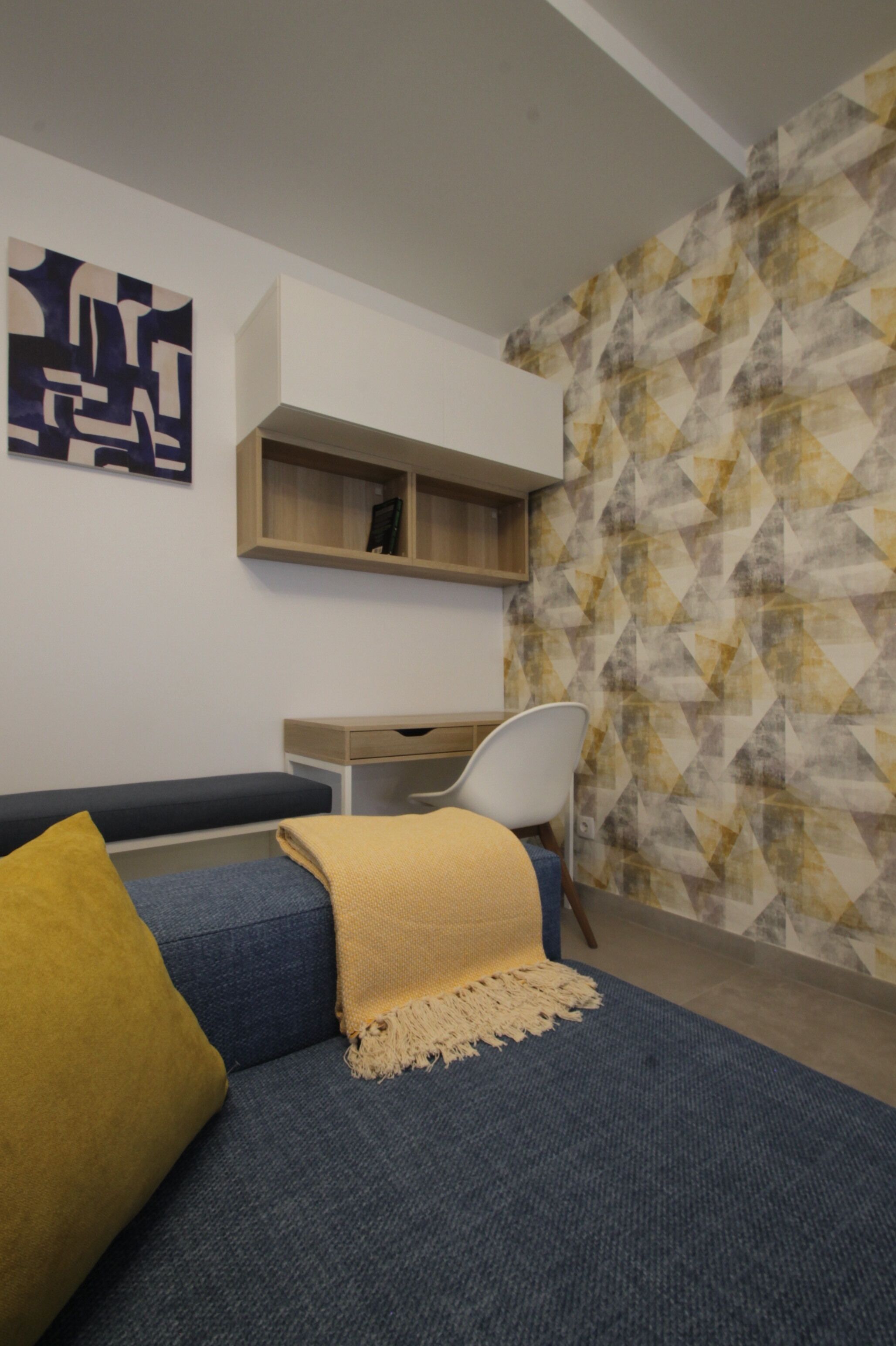 A modern living room with a blue sofa, a yellow throw, a white chair, a wooden shelf, and a geometric-patterned wall.