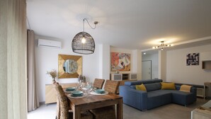A modern living room with a dining area, featuring a wooden dining table, wicker chairs, a blue sofa, and a hanging light fixture.