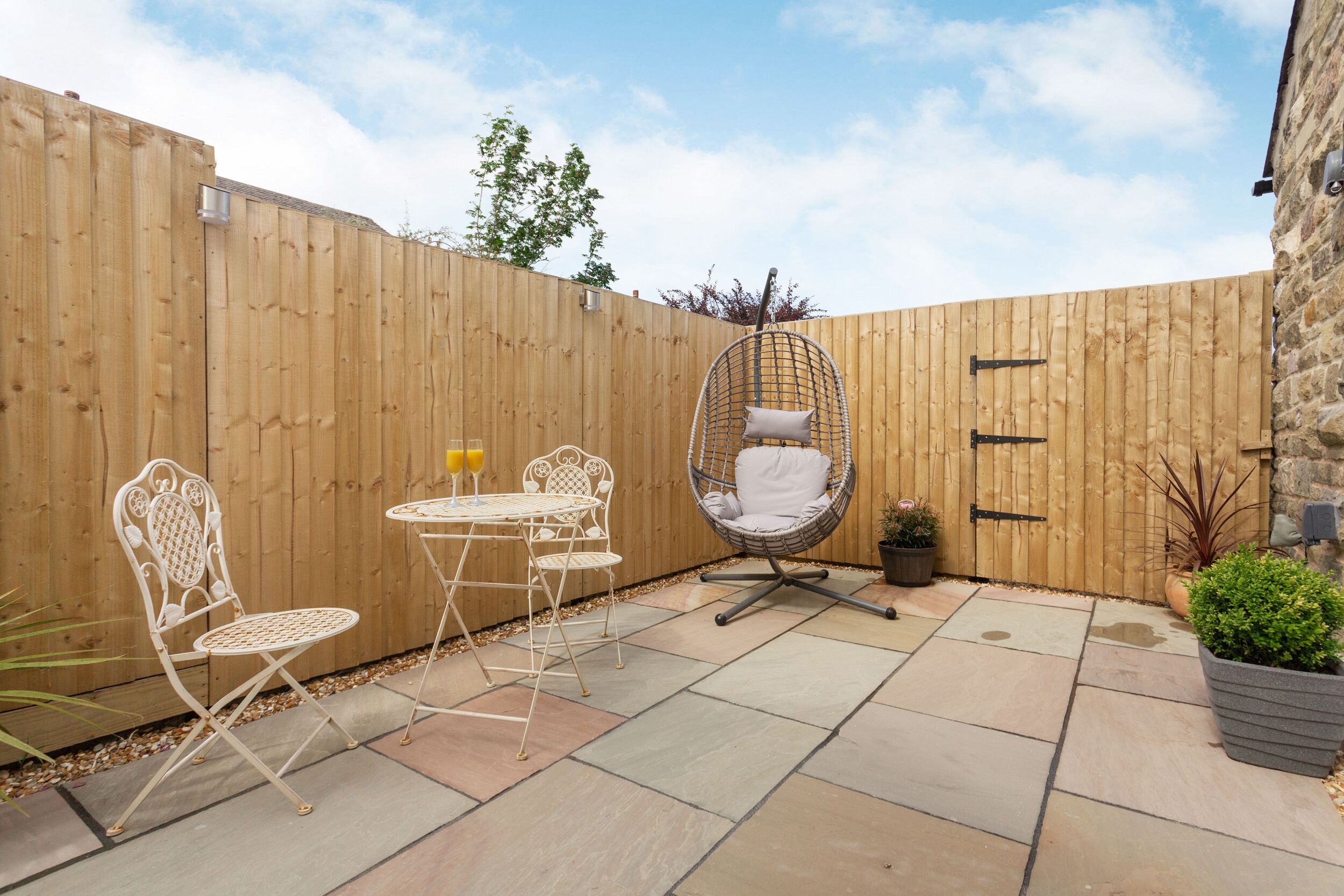 A patio with a wooden fence, a white metal table with chairs, a hanging chair, and potted plants.