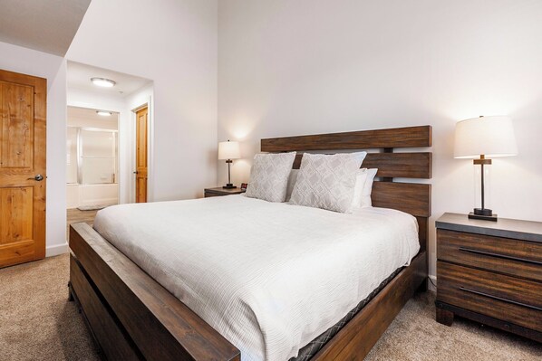 A bedroom with a wooden bed, two bedside tables with lamps, a wooden wardrobe, and a door leading to another room.