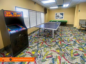 A room with a ping pong table, a racing game machine, and a green pool table.