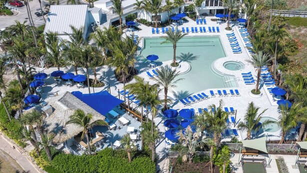 Vista aérea de un resort con piscina central, tumbonas, palmeras y una zona cubierta con sombrillas.