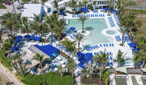 An aerial view of a resort with a central swimming pool, sun loungers, palm trees, and a covered area with umbrellas.