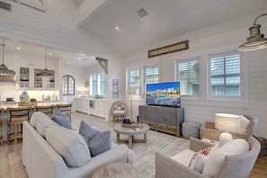 A spacious living room with a television, comfortable seating, and a kitchen area in the background.