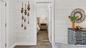 Un pasillo con piso de madera, una cama con cabecera blanca, una mesita de noche, un espejo con marco decorativo y cuadros colgados en la pared.