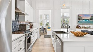 A modern kitchen with white cabinetry, a central island, and a dining area with a view. - Blue Magnolia 217 LAK (Port Aransas)