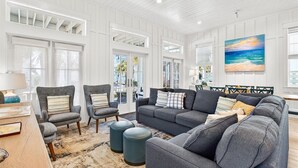 A spacious living room with a large gray sofa, two armchairs, a coffee table, and a rug. There are large windows with white shutters, a painting of a beach scene, and a view of the outdoors. - Blue Magnolia 217 LAK (Port Aransas)