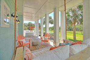 Eine überdachte Veranda mit Holzboden, zwei orangefarbene Adirondack-Stühle, eine weiße Couch mit orangefarbenen Kissen und Blick auf einen Garten mit Palmen.