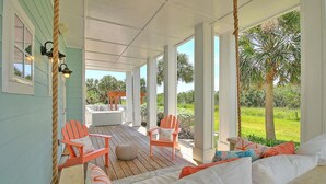A covered porch with wooden flooring, two orange Adirondack chairs, a white sofa with colorful cushions, and a view of a garden with palm trees.