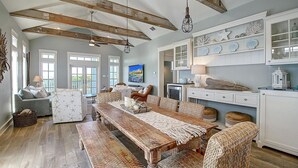 A spacious living room with a wooden dining table, wicker furniture, and a kitchen with white cabinets and decorative shells.