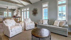 A spacious living room with a grey sofa, two white armchairs with star patterns, a round wooden coffee table, and large windows providing natural light.