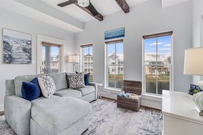 Ein modernes Wohnzimmer mit einem grauen Ecksofa, einem kleinen Beistelltisch, einem Korbwägelchen und Blick auf den Strand durch große Fenster.