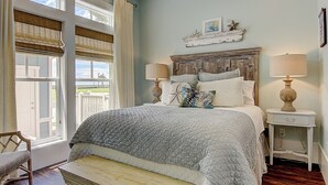A bedroom with a large bed, two bedside lamps, a wooden bench, and a view of the outdoors through a window with blinds. - Beach Crush TC 7102 (Port Aransas)