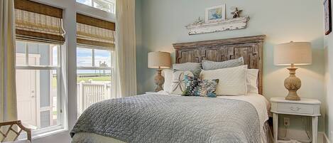 A bedroom with a large bed, two bedside lamps, a wooden bench, and a view of the outdoors through a window with blinds.
