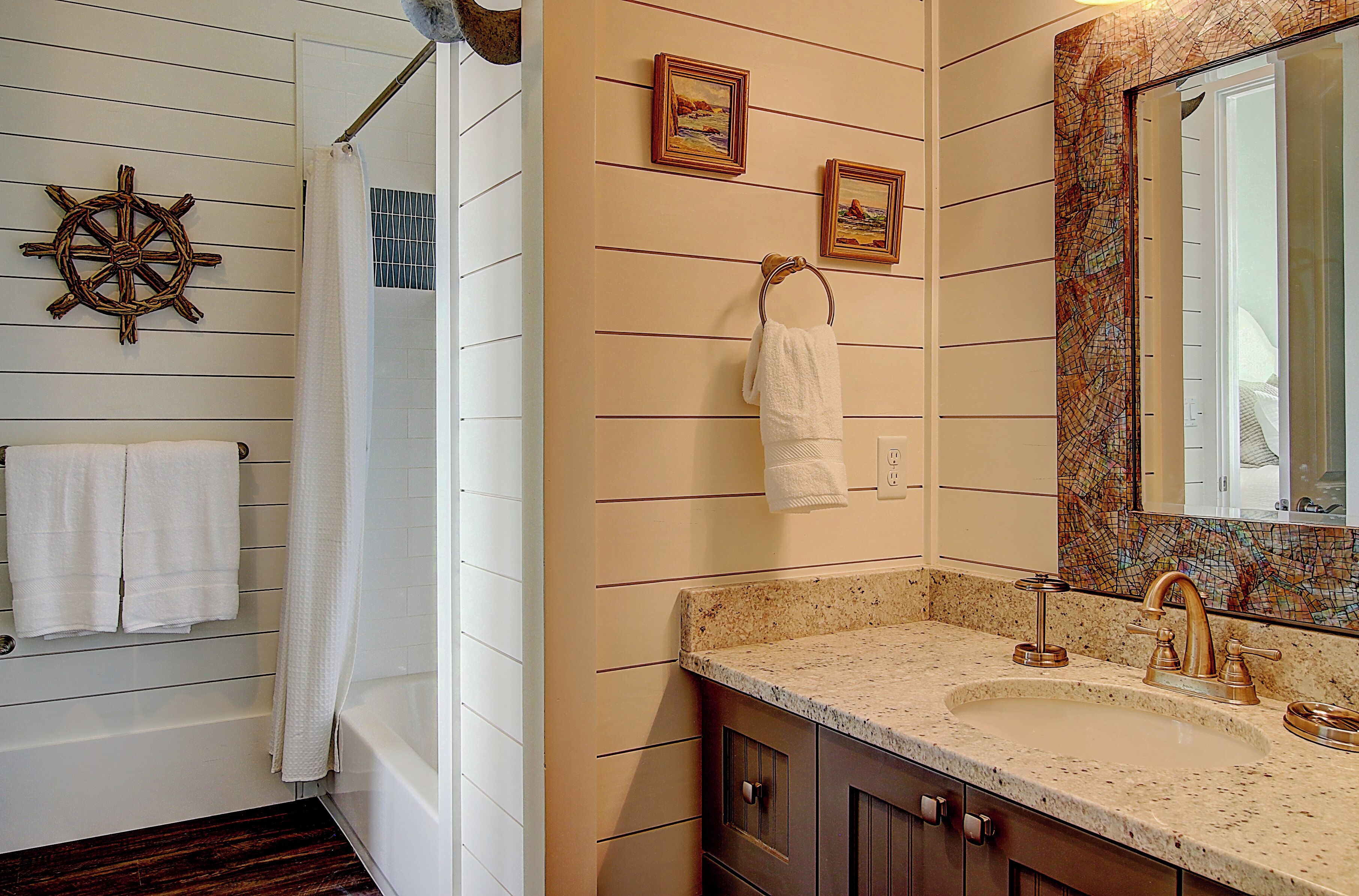Une salle de bain avec une baignoire blanche, un lavabo avec un meuble-vasque, un miroir avec un décor en mosaïque et une décoration murale représentant un gouvernail de bateau.