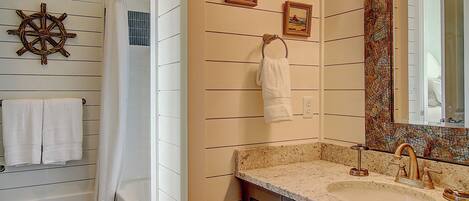 Une salle de bain avec une baignoire blanche, un lavabo avec un meuble-vasque, un miroir avec un décor en mosaïque et une décoration murale représentant un gouvernail de bateau.